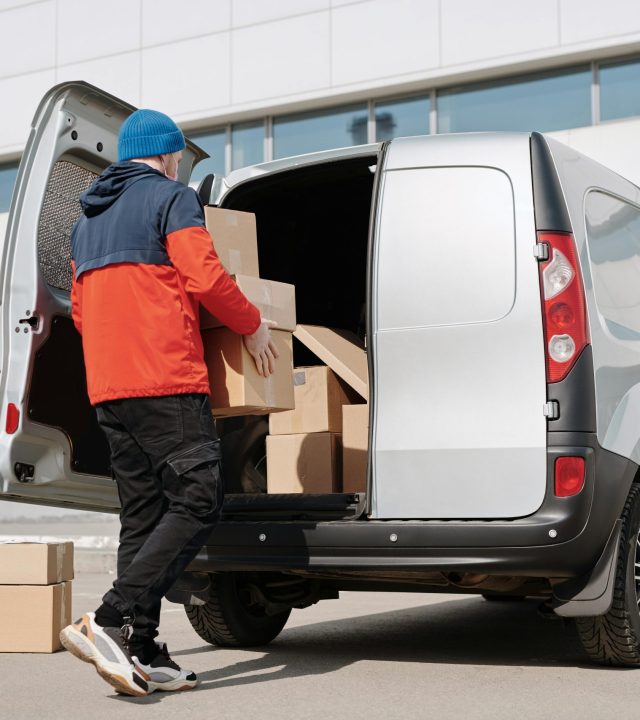 Man in colorful jacket loading cardboard boxes into a van outside an office building.
