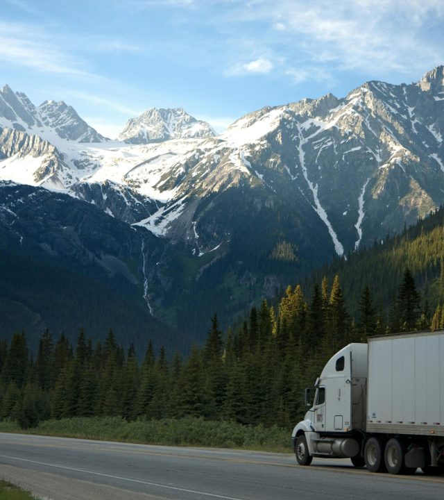 A semi-truck travels along a highway with snow-capped mountains in the background.